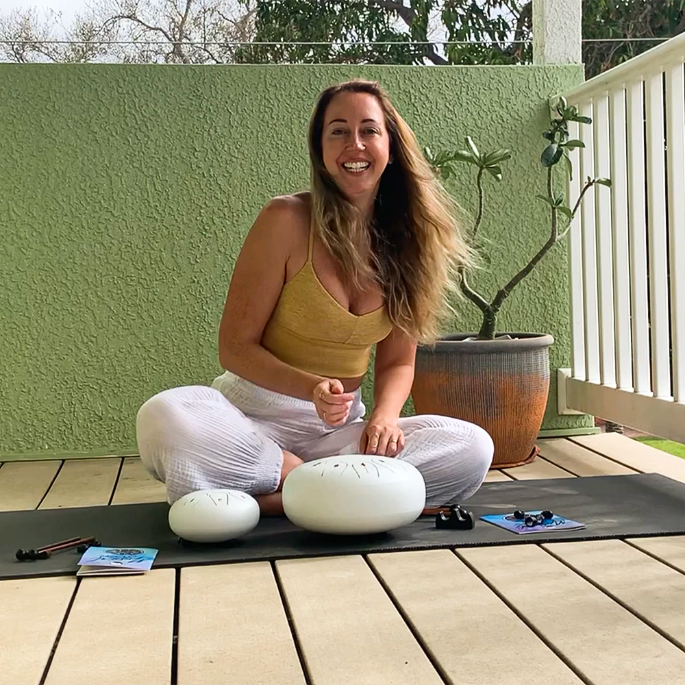 Woman sitting cross-legged on a yoga mat outdoors with two white steel tongue drums