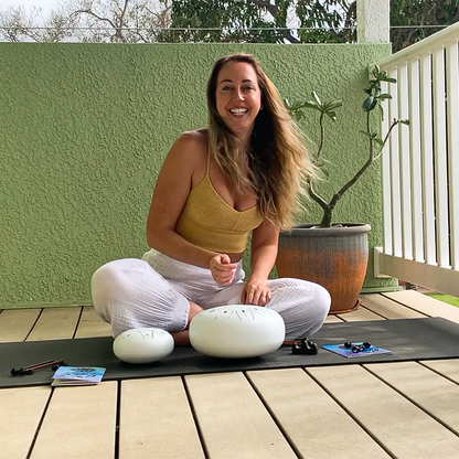 Woman sitting cross-legged on a yoga mat outdoors with two white steel tongue drums