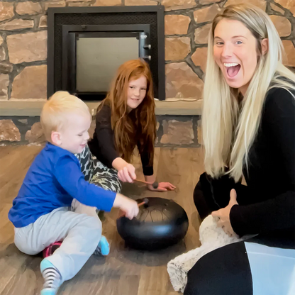Mother and two young children playing a black steel tongue drum together on the floor