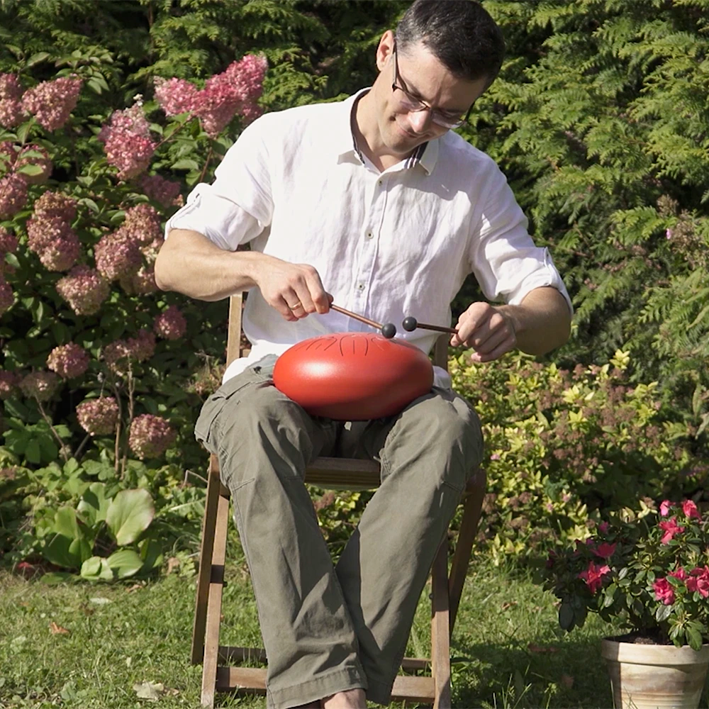 Man sitting on a chair in a garden playing a steel tongue drum with mallets